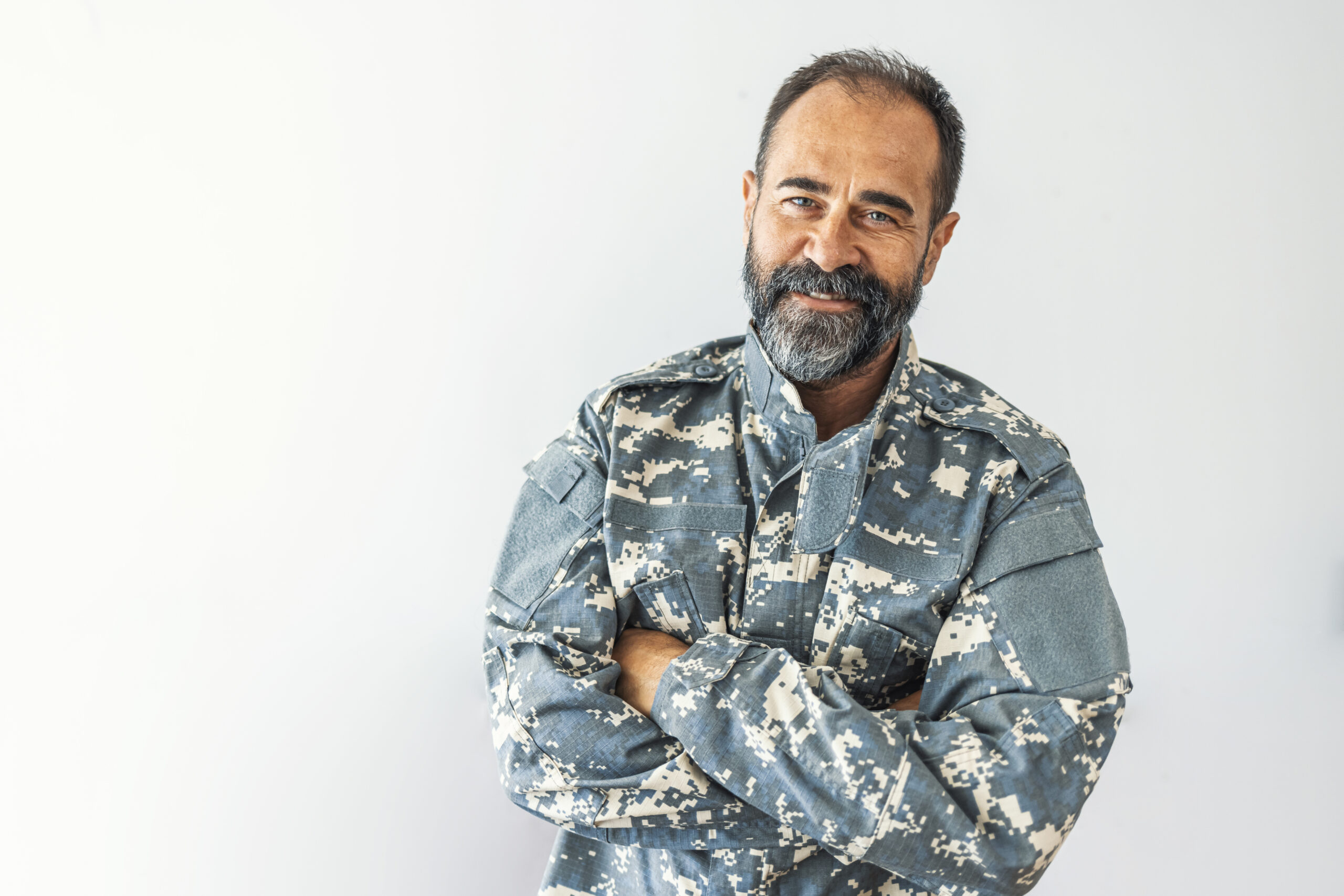 Veteran with beard in camouflage uniform standing with arms crossed.