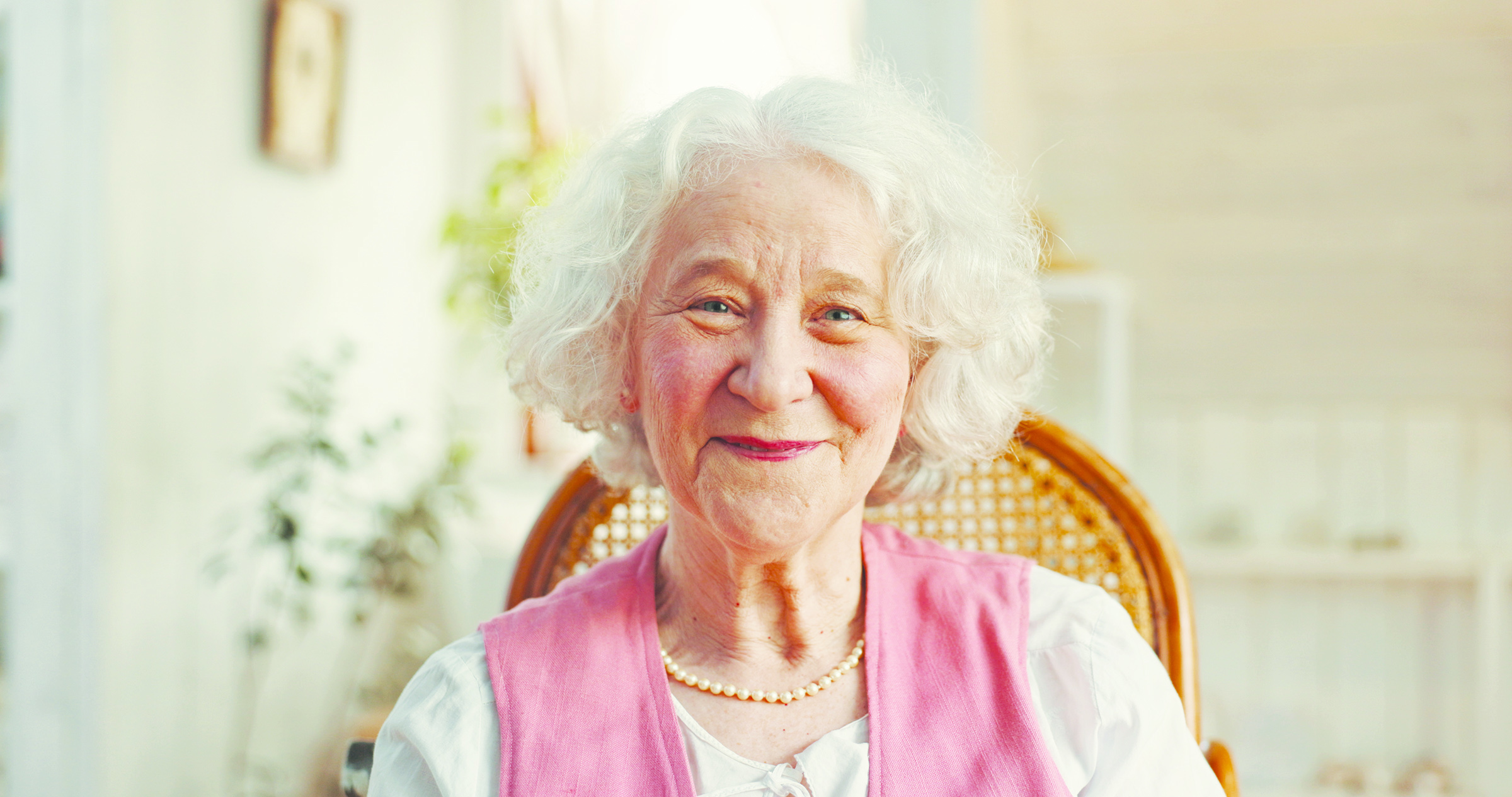 Smiling elderly woman with white hair sitting in a bright, peaceful home environment.