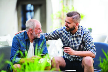 A young man and his elderly father sharing coffee outdoors, engaged in a conversation about end of life planning.